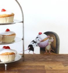 two cupcakes with white frosting and red cherries on them are in front of a mouse figurine