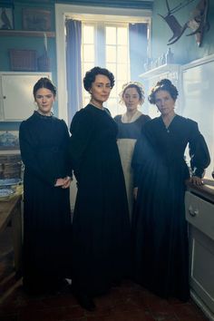 three women in long black dresses stand next to each other near an oven and sink