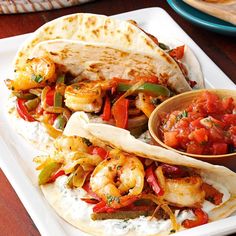 two tacos with shrimp, peppers and onions on a plate next to tortillas