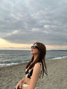 a woman standing on top of a sandy beach next to the ocean holding a white purse