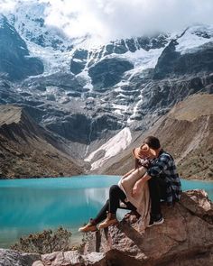 a man and woman sitting on top of a rock next to a lake