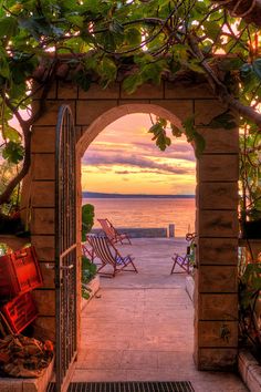 an archway leading to the beach with lawn chairs on either side and sunset in the background