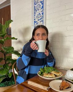 a woman sitting at a table drinking from a coffee cup with food on the side