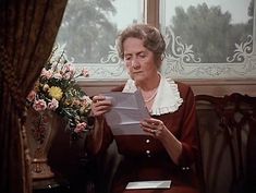 an older woman sitting on a couch reading a book in front of a vase with flowers