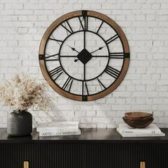 a large clock mounted to the side of a white brick wall next to a vase with dried flowers