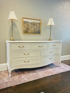 a white dresser sitting on top of a hard wood floor next to a lamp and painting