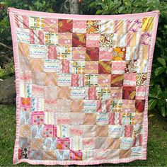 a patchwork quilt hanging from a tree in front of some bushes and flowers on the grass