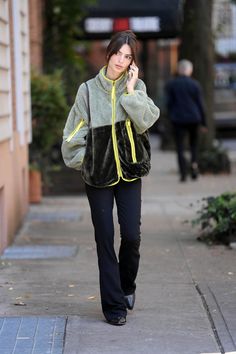 a woman is walking down the sidewalk talking on her cell phone while wearing a green and black jacket