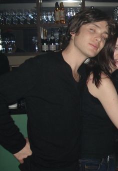 a man and woman standing next to each other in front of a bar with bottles on the shelves