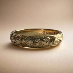 a gold wedding band with flowers and leaves engraved on the side, sitting on a plain surface