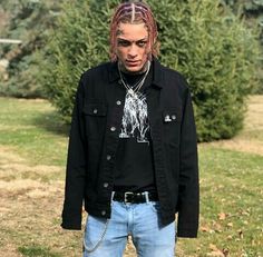 a young man with dreadlocks standing in front of some bushes and trees wearing a black jacket