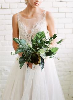 a woman in a wedding dress holding a bouquet with greenery and leaves on it