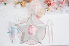 a pink and white christmas table setting with silver star, candle holder, utensils