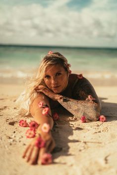 a woman laying on top of a sandy beach next to the ocean with flowers in her hair