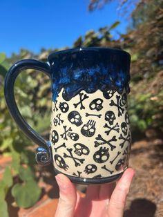 a hand holding up a blue and white coffee mug with skulls on it, in front of cacti