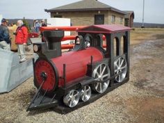 a toy train sitting on top of a gravel field next to a building with people standing around it