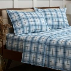 a bed with blue and white plaid sheets on it, next to a vase filled with dried flowers