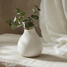 a white vase filled with green leaves on top of a table next to a curtain