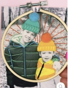 a hand holding up a small embroidered picture with two boys wearing hats on it and one boy in the background