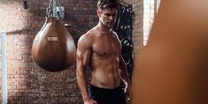 a shirtless man standing next to a punching bag in a brick wall - mounted gym