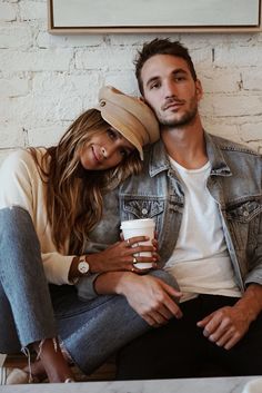 a man and woman sitting next to each other holding coffee cups in front of a brick wall