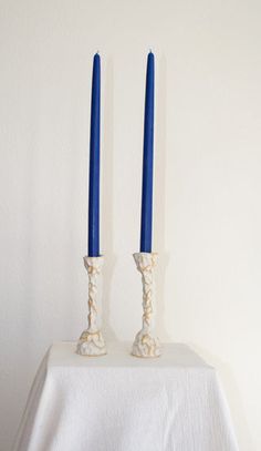 two blue candles sitting on top of a white table cloth next to each other in front of a white wall