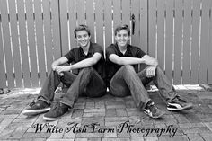 two young men sitting on the ground in front of a fence with their arms crossed