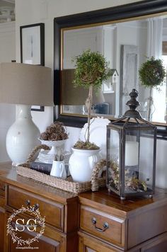 a table topped with white vases filled with plants and a mirror behind it on top of a wooden dresser