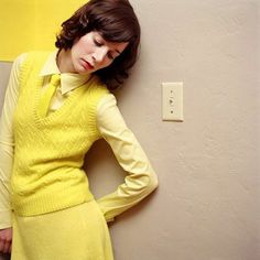 a woman leaning against a wall with her hands on her hips, wearing a yellow dress