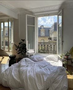 an unmade bed with white sheets and pillows in a room next to two windows