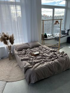 an unmade bed sitting in front of a window next to a rug and vase