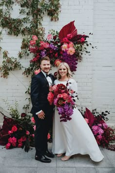 a bride and groom standing in front of a wall with flowers on it, posing for the camera