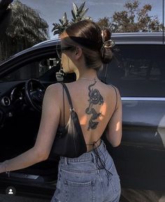 a woman with a dragon tattoo on her back standing next to a car and holding the door open