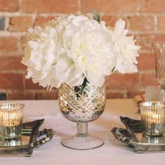 white flowers are in a silver vase on a table with gold dishes and glasses around it