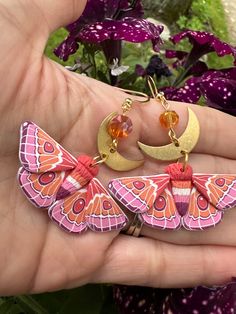 a hand holding three butterfly shaped earrings in front of purple flowers with gold chains on them