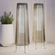 two tall metal vases sitting next to each other on a white tile floor with a plant in the middle