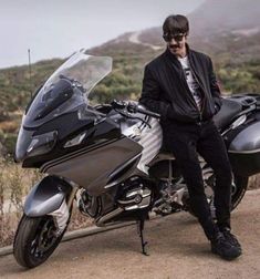 a man sitting on the back of a motorcycle next to a dirt road with mountains in the background