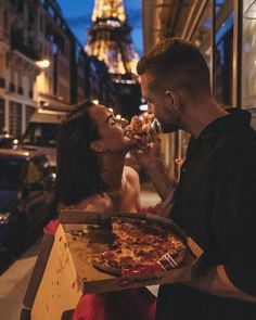 a man and woman eating pizza in front of the eiffel tower at night