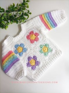 a crocheted baby sweater with flowers on it and a plant in the background