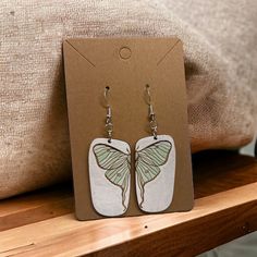 Luna moth design engraved in birch wood, each earring featuring one side of the luna moth.  Hand painted in soft blues and greens, with white background. Earrings are an organic rectangular shape with more rounded edges, and feature hypoallergenic silver plated hooks. Moth Earrings, Luna Moth