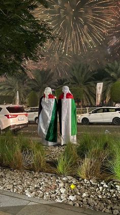 two people dressed in white and green standing next to each other with fireworks behind them