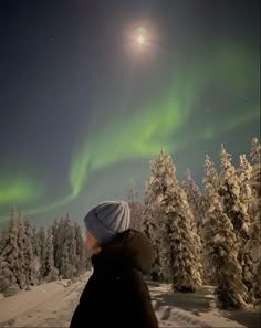 a person standing in the snow looking up at an aurora bore over trees and bushes