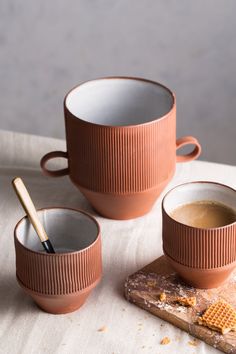 three coffee cups with spoons on top of a cutting board and some crackers