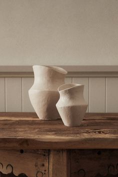 two white vases sitting on top of a wooden table in front of a wall