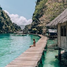 a person sitting on a dock in the water next to some mountains and houses with thatched roofs