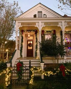 a white house decorated for christmas with lights and wreaths on the front porch, surrounded by greenery