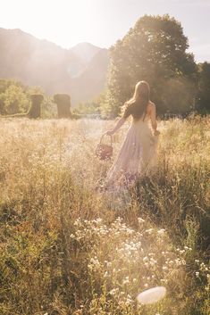 a woman in a dress walking through a field with a basket on her back and mountains in the background