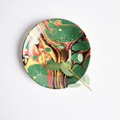 a green and red plate with leaves on it sitting on a white surface next to a plant