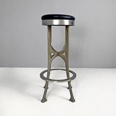 a metal stool with a black seat on a white tableclothed floor in front of a gray wall