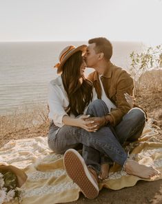 a man and woman sitting on a blanket near the ocean kissing while holding each other